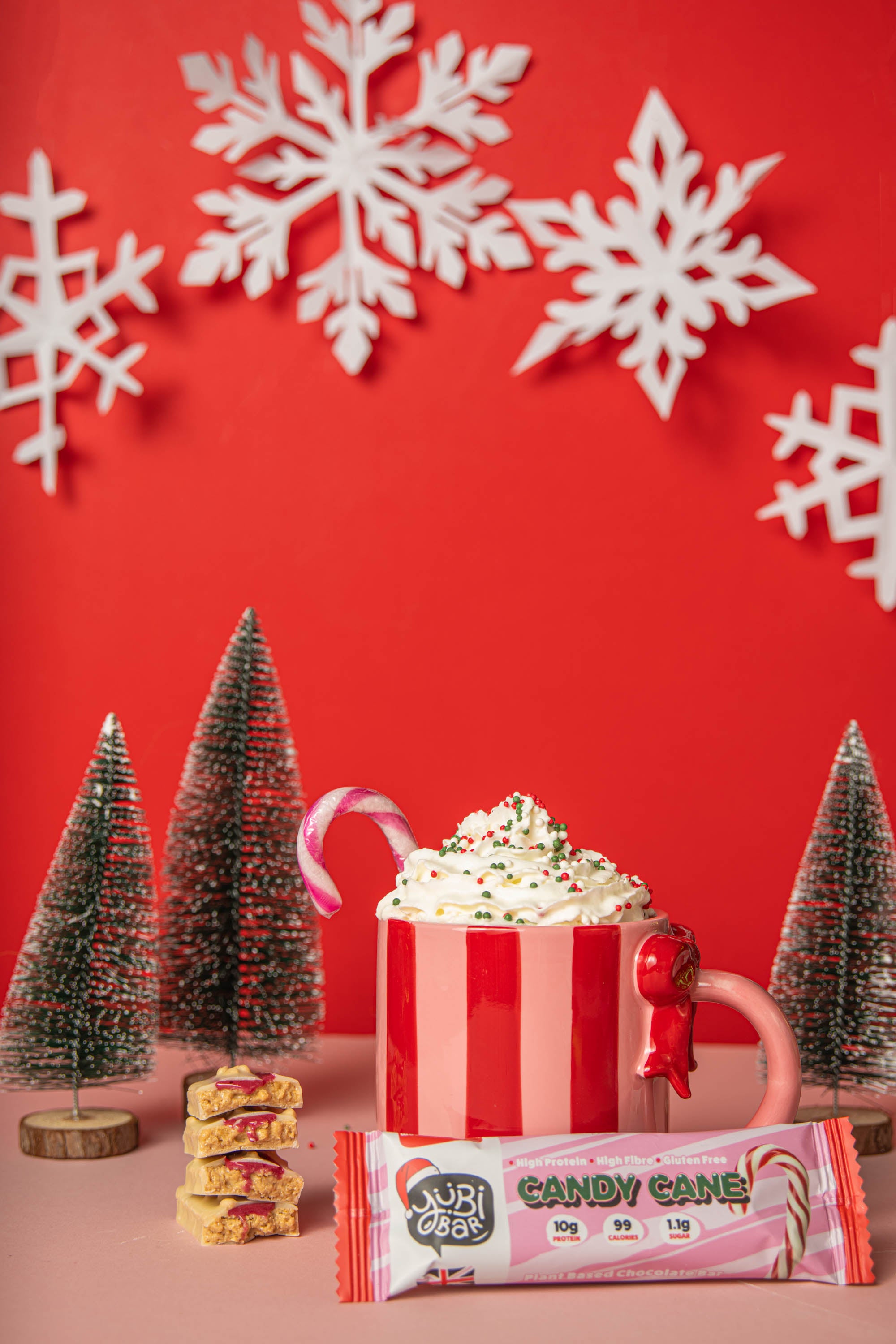YuBi Candy Cane Christmas Protein Bar and Hot chocolate with whipped cream and a candy cane in a striped mug on a red background with snowflakes and Christmas trees.